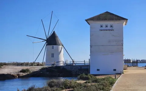Altes Windrad und ein weißes Gebäude am Mar Menor, mit Wasser und Vegetation im Vordergrund.