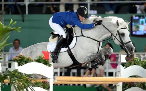 Une cavalière en tenue bleue et blanche saute un obstacle avec son cheval gris. Le cheval, nommé Levisto Z, montre une puissance impressionnante.