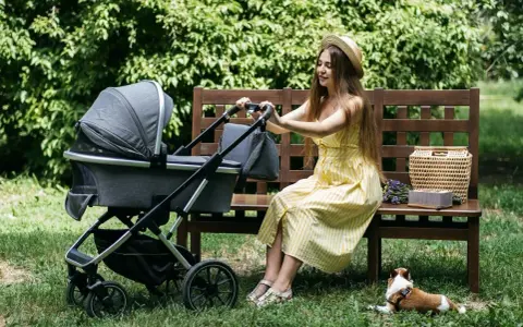 Kobieta w żółtej sukience siedzi na ławce obok wózka Kinderkraft. Piesek leży na trawie.