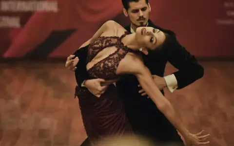 Para tańczy tango, jeden z rodzajów tańca towarzyskiego. Kobieta w bordowej sukni i mężczyzna w czarnym fraku wykonują skomplikowaną figurę.