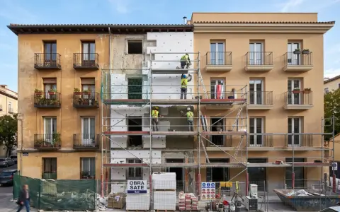 Obreros instalan aislamiento térmico en fachada de edificio. Se ve el **sate lana de roca** en bloques blancos, andamios y materiales de construcción.