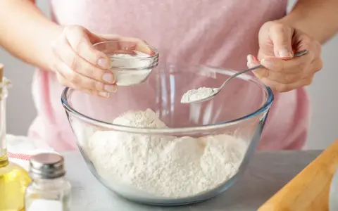 Kobieta dodaje do mąki w szklanej misce biały proszek, kluczowy składnik proszku do pieczenia.
