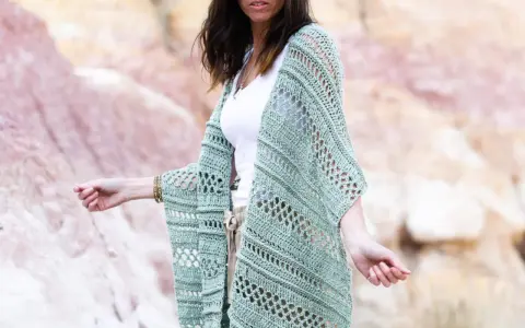Mujer con gafas de sol luciendo un chaleco de **mantones de crochet** verde menta, con un diseño calado y mangas anchas, sobre una camiseta blanca y pantalones beige.