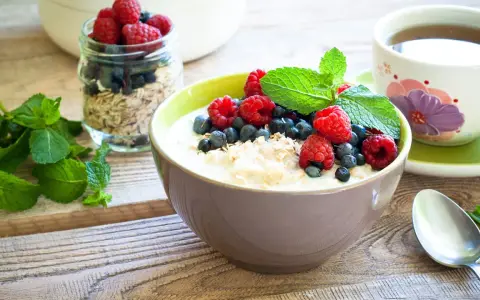 Schüssel mit Haferflocken mit Kefir, garniert mit Himbeeren, Blaubeeren und Minze. Daneben ein Glas Müsli und eine Tasse Tee. Gesundes Frühstück.