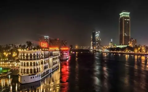 Beleuchtete Nilschiffe und moderne Hochhäuser spiegeln sich im Wasser. Ein magischer Anblick von Kairo bei Nacht.