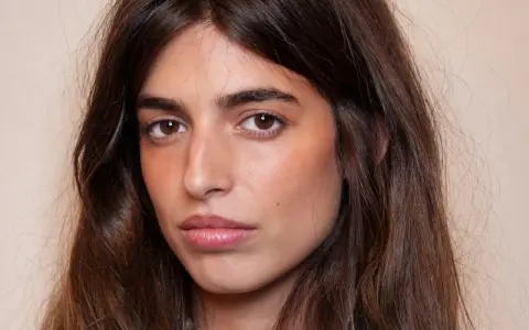 Portrait d'une jeune femme aux longs cheveux bruns. Son maquillage met en valeur ses yeux et ses lèvres, avec un soin particulier apporté à son sourcil poil à poil.