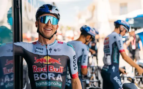 Cyclist smiles on a vuelta rest day, wearing a BORA-hansgrohe kit with Red Bull logos. Other team members and bikes are visible.