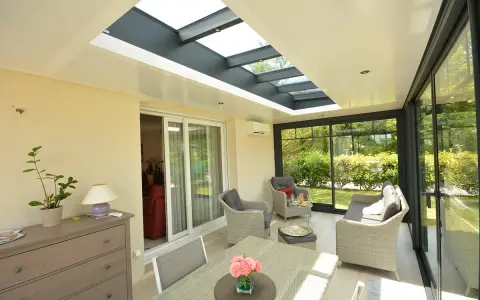 Une **veranda qui s'ouvre entièrement** sur un jardin verdoyant. Mobilier confortable, table et chaises pour profiter du soleil.