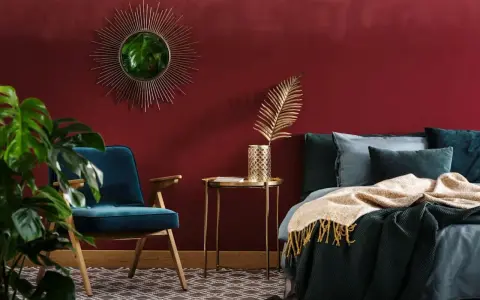 Habitación con pared de pintura a la tiza roja, sillón azul, cama con mantas y cojines, espejo sol y planta.