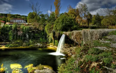 Cascadas en España: una casa rural se asoma entre la vegetación exuberante junto a una cascada que cae en un estanque de aguas verdes.