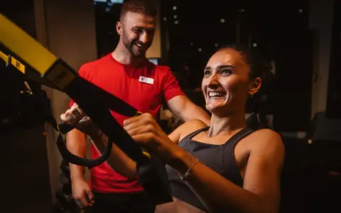 Frau lacht und trainiert mit TRX-Bändern, ihr Trainer lächelt. Sie will schnell wieder fit werden.