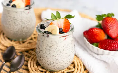 Zwei Gläser mit cremigem Chia Pudding, garniert mit Erdbeeren, Bananen und Blaubeeren. Daneben eine Schale frischer Erdbeeren.