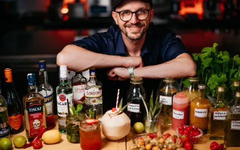 Uśmiechnięty mężczyzna w czapce i okularach za ladą pełną alkoholi i owoców. To przyszłość: drink bar zamiast barmana.