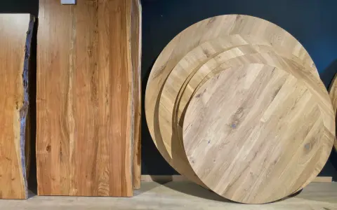 Plusieurs plateaux de table ronde en bois, empilés, illustrent la définition d'une table ronde. Des planches de bois brut sont aussi visibles.