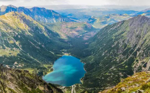 Widok na Morskie Oko z góry. W dole błękitna tafla jeziora otoczona lasem i górami. Kamera uchwyciła parking w oddali.