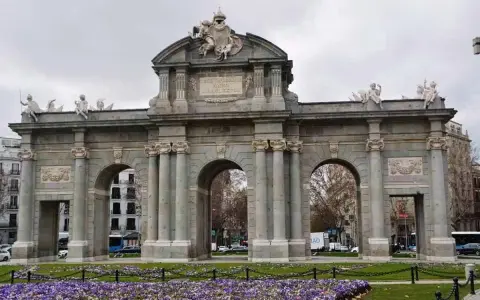 La majestuosa Puerta de Alcalá se alza imponente, con sus arcos y esculturas, rodeada de un jardín de flores moradas.