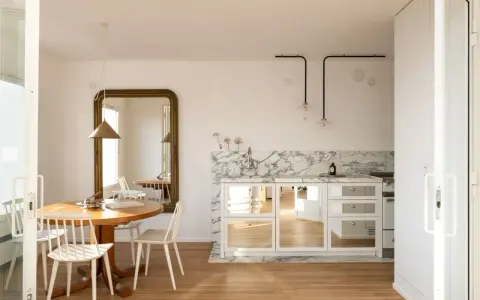 Comedores pequeños con encanto, con mesa redonda de madera, sillas blancas y un gran espejo dorado. La cocina integrada tiene encimera de mármol y armarios con puertas de espejo.