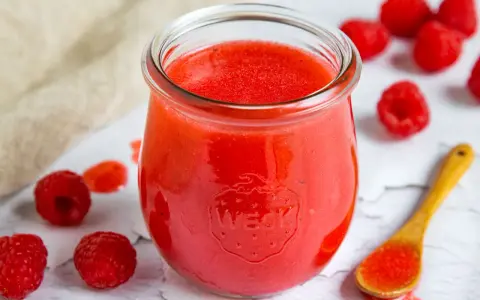 Ein Glas mit leuchtend roter Himbeer-Vinaigrette, umgeben von frischen Himbeeren und einem Holzlöffel mit etwas Vinaigrette.