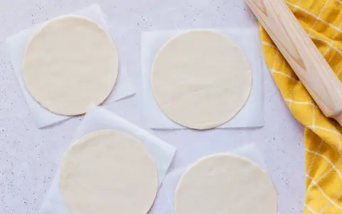 Cuatro discos de masa empanada listos para rellenar, junto a un rodillo y un paño amarillo.