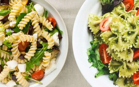 Due ciotole di pasta fredda gourmet: una con fusilli, melanzane, pomodorini e mozzarella; l'altra con farfalle al pesto e pomodorini.