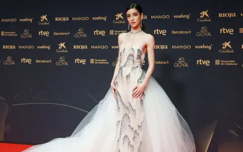 Una joven desfila en la goya alfombra roja con un vestido de gala blanco con detalles plateados y una voluminosa cola de tul.