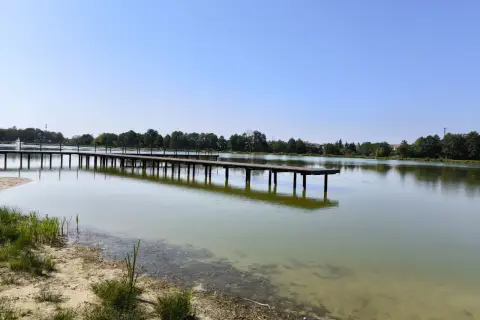 Drewniane molo w Krasnosielcu w letni, słoneczny dzień. Woda spokojna, odbija niebo i drzewa na horyzoncie.