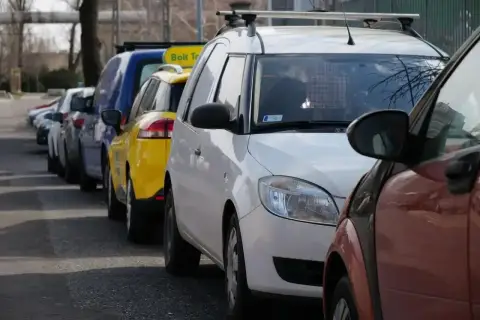 Rząd samochodów, w tym żółty Bolt Taxi, zaparkowany na chodniku.