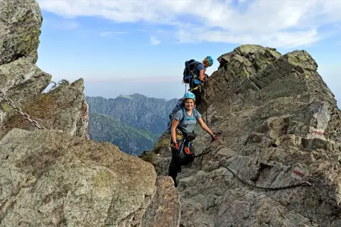 Dwoje turystów wspina się po skalistych tatry wysokie szlaki, korzystając z łańcuchów. Widok na góry i błękitne niebo.