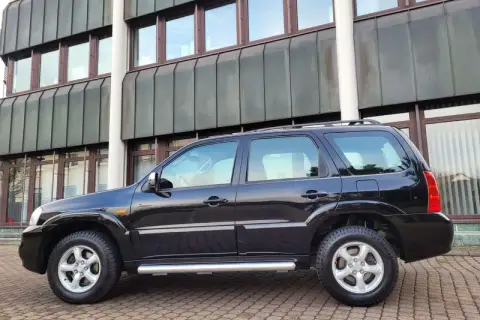 Czarny Mazda Tribute stoi na brukowanym podjeździe przed nowoczesnym budynkiem.