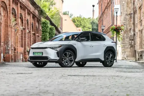 Biały Toyota bz4x na brukowanej uliczce starego miasta, otoczony ceglanymi budynkami i zielenią.