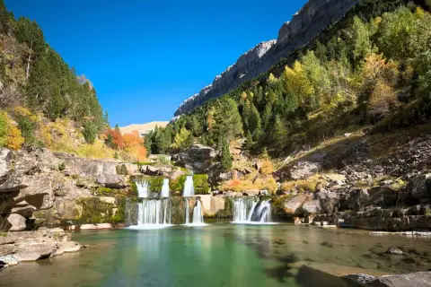 Cascada en un valle montañoso, ideal para rutas senderismo España. El agua verde turquesa fluye entre rocas y árboles otoñales.