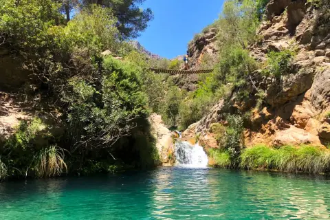 Un puente colgante cruza un **río verde** en Granada, con una cascada y vegetación exuberante.