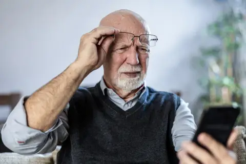 Starszy mężczyzna z siwą brodą i łysiną, poprawiając okulary, z trudem skupia wzrok na ekranie telefonu. Czy to początki demencji?