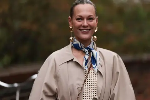 Uśmiechnięta kobieta w beżowym płaszczu i stylowej apaszce z niebieskimi wzorami. Inspiracja, jak nosić apaszkę, by dodać elegancji.