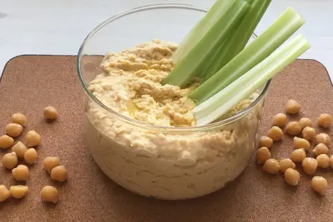 Domowy hummus przepis: kremowa pasta z ciecierzycy w szklanej misce, podana z łodygami selera i ziarnami ciecierzycy.