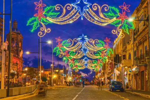 Malta w grudniu rozświetlona świątecznymi iluminacjami. Ulica ozdobiona girlandami z gwiazd i kwiatów, w tle kościół i sklepy.