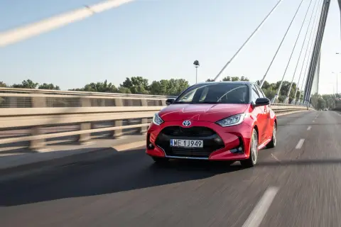 Czerwony Toyota Yaris Hybrid pędzi po moście w słoneczny dzień.