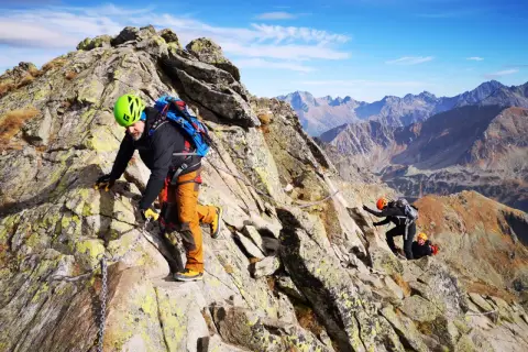 Wspinacze pokonują skalistą grań, a jeden z nich, w zielonym kasku, zmierza w stronę szczytu. Widok na Tatry zapiera dech w piersiach, a świnica przez zawrat wydaje się być na wyciągnięcie ręki.