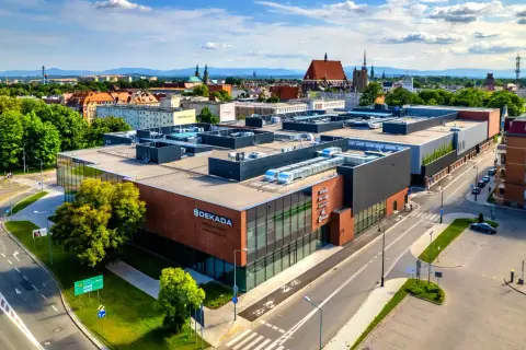 Nowoczesny budynek usługowy "Dekada" z przeszkloną fasadą i ceglanymi elementami, otoczony zielenią i drogami.