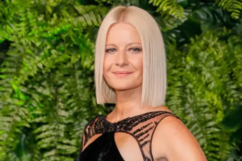 Małgorzata Kożuchowska w czarnej sukni z ozdobnym dekoltem na tle zielonych liści.