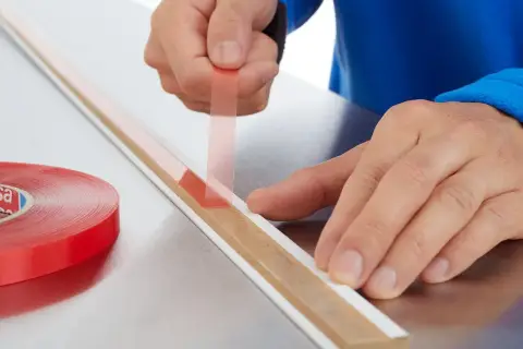 Ręce przykładają cienką taśmę dwustronną do listwy. Obok leży rolka czerwonej taśmy.