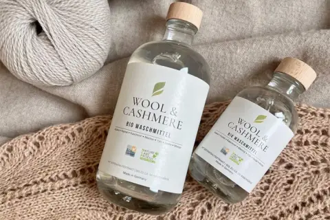 Zwei Flaschen "Wool & Cashmere" Bio-Waschmittel neben Garnknäueln und Strickwaren. Perfekt für die sanfte **Wolle waschen mit Shampoo**.