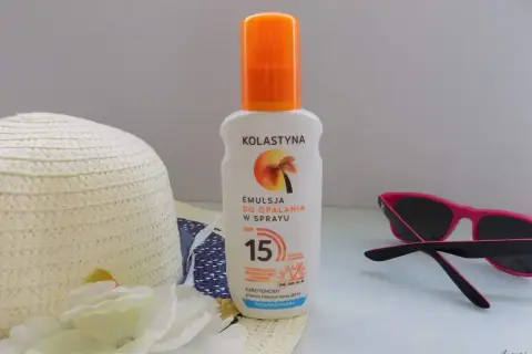 Emulsja do opalania w sprayu Kolastyna SPF 15 z karotenoidami, wodoodporna. Obok kapelusz i okulary przeciwsłoneczne.