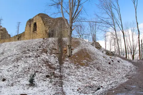 Ruiny zamku Lanckorona na wzgórzu pokrytym śniegiem. Kamienne mury wznoszą się na tle błękitnego nieba, a wokół rosną nagie drzewa.