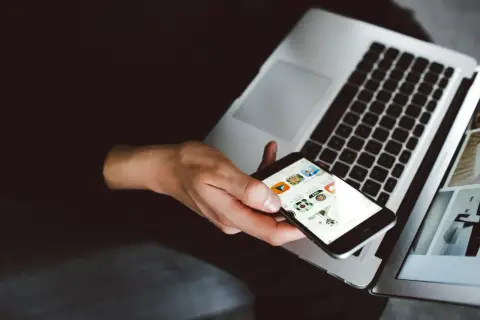 Dłoń trzyma iPhone'a z widocznymi ikonami aplikacji, obok otwartego laptopa. To idealny moment na **backup iPhone'a**.