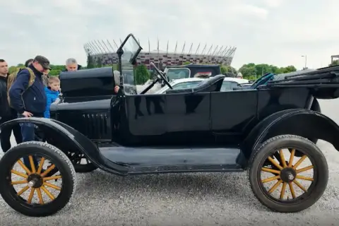 Czarny, zabytkowy Ford T z drewnianymi szprychami kół, obok którego stoją ludzie. W tle widać stadion.
