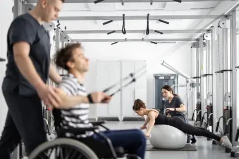 Rehabilitacja metodą Bobath: mężczyzna na wózku ćwiczy z fizjoterapeutą, kobieta na piłce z hantlami pod okiem terapeutki.