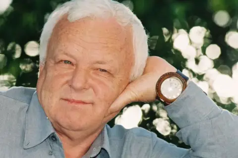 Uśmiechnięty Marian Opaniak, z siwymi włosami i niebieskimi oczami, opiera głowę na dłoni. Zegarek na jego nadgarstku wskazuje godzinę.