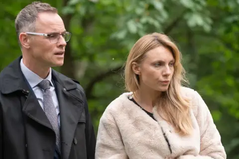 Adam i Ewa obsada: mężczyzna w okularach i płaszczu oraz kobieta w futrzanej kurtce na tle zieleni.