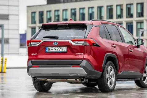 Czerwony, używany Toyota RAV4 Hybrid z salonu, zaparkowany na mokrym parkingu przed nowoczesnym budynkiem.